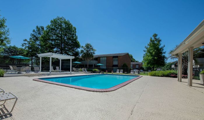 Inviting outdoor pool with elegant poolside seating, surrounded by lush greenery and charming apartment view under a vibrant blue sky.