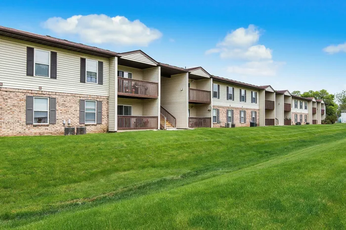 Charming apartment complex with brick and siding exterior, featuring private balconies and lush green lawns under a bright blue sky.