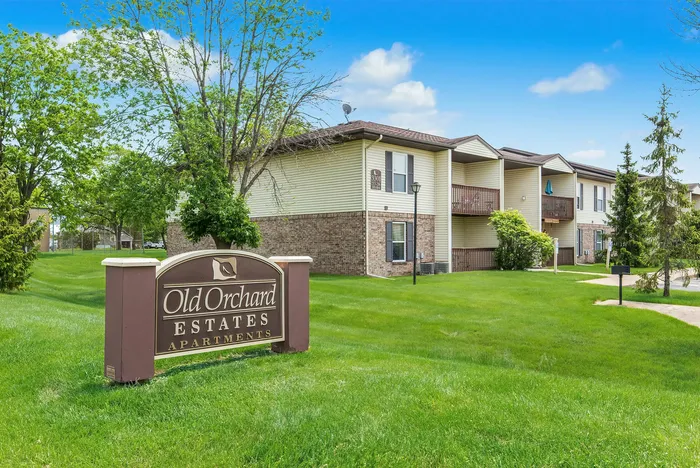 Charming Old Orchard Estates Apartments with lush green lawns and inviting balconies surrounded by vibrant trees and blue skies.