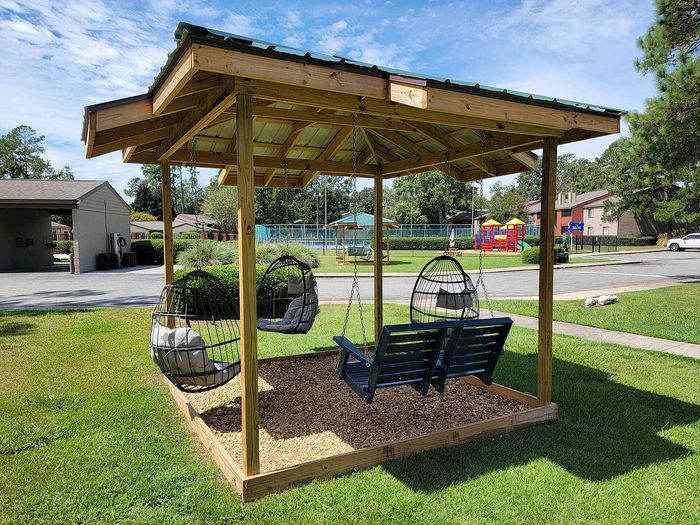 Charming outdoor gazebo with cozy hanging chairs and a swing, set in a lush green garden near a playground.