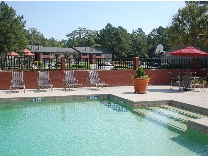 Inviting community pool area featuring comfortable lounge chairs and vibrant red umbrellas, surrounded by lush greenery and stylish apartments.