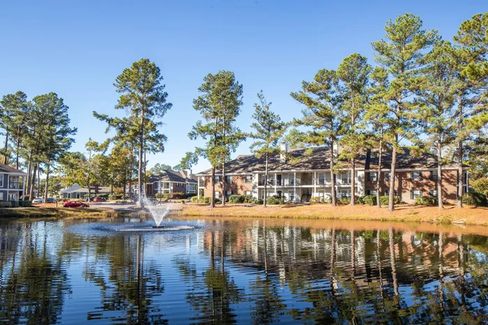 Charming apartment community nestled among tall pine trees with a serene pond and fountain reflecting the blue sky.