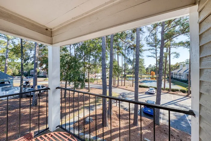 Charming balcony view overlooking a serene landscape with tall pine trees, spacious parking, and a distant pond, perfect for peaceful relaxation.