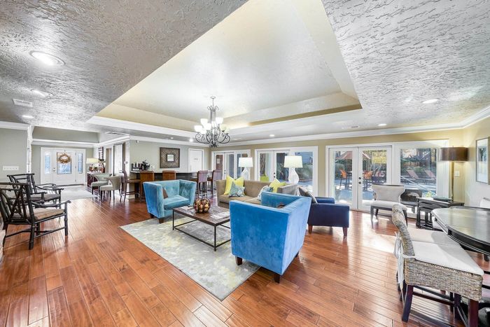 Elegant and spacious living area with bright lighting, modern blue velvet chairs, hardwood floors, and a stunning view of the pool area through large glass doors.