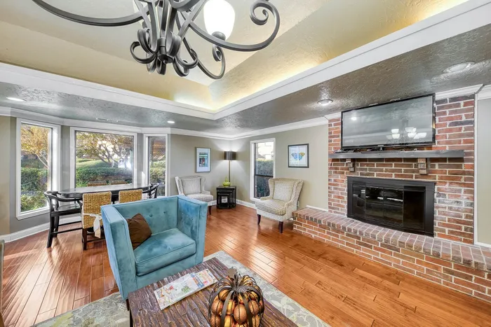 Charming living room with elegant chandelier, cozy brick fireplace, and large windows revealing lush greenery.