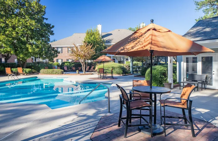 Charming outdoor pool area with elegant seating under vibrant umbrellas, surrounded by lush greenery and a beautiful brick apartment building.
