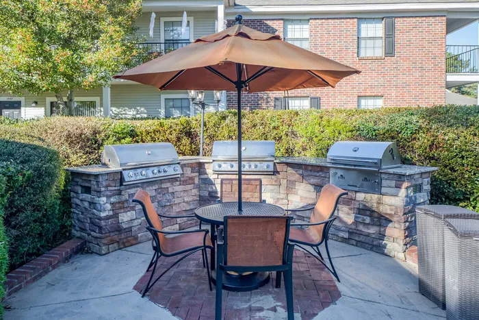 Charming outdoor grilling area with modern brickwork, featuring three stainless steel BBQ grills, a stylish patio table, and comfortable seating under a wide sunshade umbrella, ideal for entertaining and relaxation.