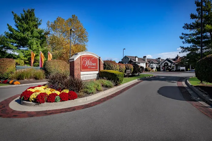 Inviting entrance of Willows of Coventry featuring vibrant landscaping, colorful flowers, and tree-lined pathways under a clear blue sky.