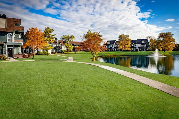 Charming lakeside community with well-manicured lawns, vibrant autumn foliage, and elegant homes surrounded by serene waters and a picturesque fountain.