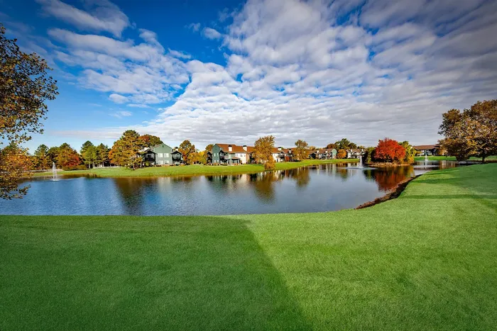 Picturesque view of elegant lakeside homes surrounded by lush greenery and vibrant autumn trees, under a clear blue sky.