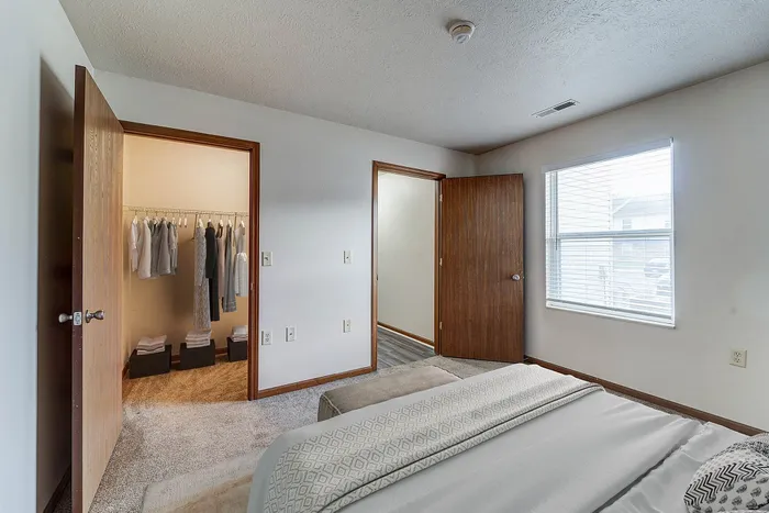 Bright bedroom with plush carpeting, featuring a cozy bed, ample natural light, and an open walk-in closet with organized storage.