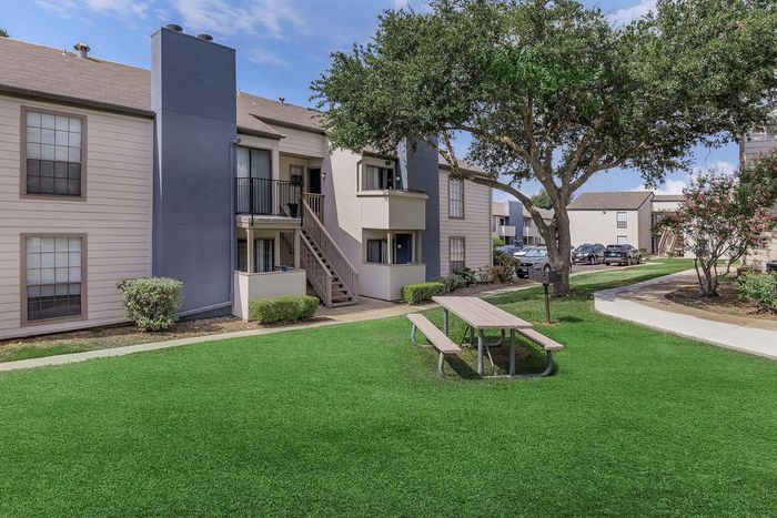 Charming apartment complex featuring lush green grounds, mature trees, and welcoming outdoor seating area.