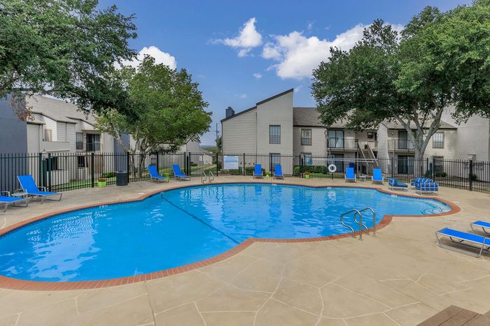 Inviting residential swimming pool area with modern lounge chairs, surrounded by lush greenery and stylish apartment buildings under a clear blue sky.
