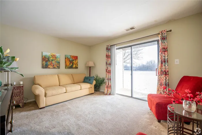 Bright living room with a large beige sofa, vibrant red chairs, and a beautiful view through sliding glass doors. Elegant decor with colorful artwork and a cozy atmosphere.