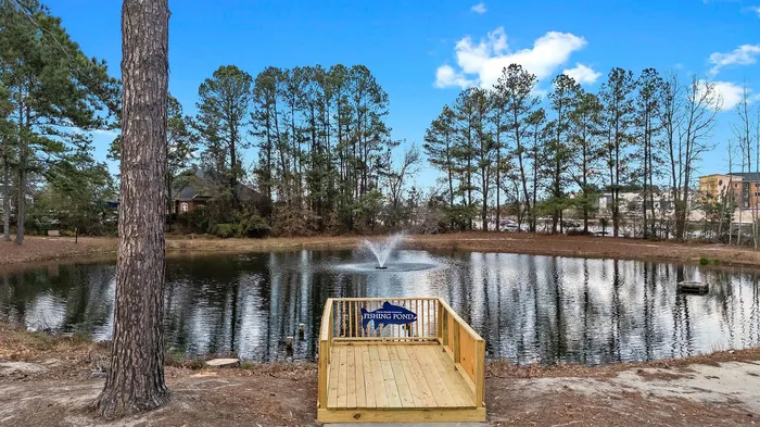 Serene pond view with a charming wooden deck and tranquil fountain, surrounded by lush pine trees and blue sky.
