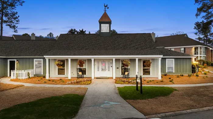 Charming clubhouse exterior at dusk with inviting rocking chairs and rustic weather vane, set amidst a picturesque landscaped garden.