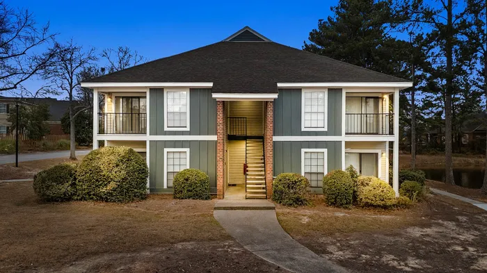 Charming two-story apartment building surrounded by lush greenery and mature trees, offering inviting balconies and a warm, welcoming entrance.