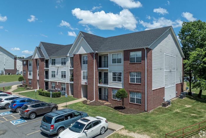 Modern three-story brick apartment building with charming balconies set in a lush green neighborhood; ample parking available under a clear blue sky.