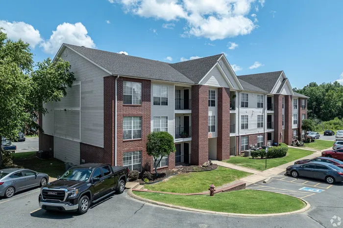 Modern apartment building with red brick facade, lush landscaping, and ample parking under a clear blue sky.