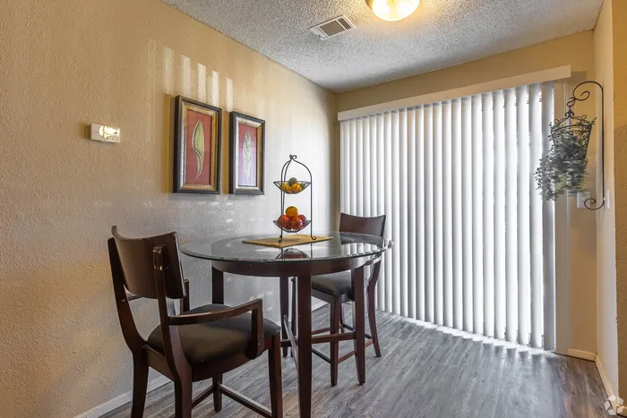 Inviting dining area with elegant round glass table, cozy chairs, and modern artwork, illuminated by natural light through vertical blinds.