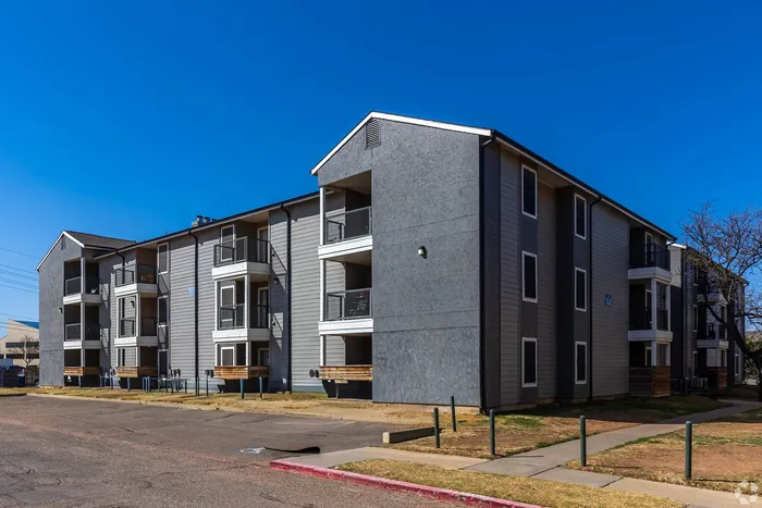 Modern apartment building with three floors, featuring private balconies and a sleek gray exterior under a clear blue sky.
