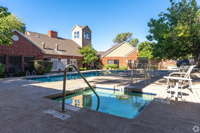 Charming apartment complex featuring a sparkling pool and inviting outdoor seating area, set against a backdrop of rustic brick architecture and lush greenery.