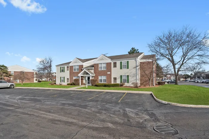 Charming two-story apartment building with brick facade and green shutters, surrounded by manicured lawns and ample parking space.