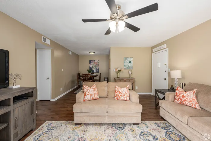Inviting living room with elegant wall art, plush beige sofas with vibrant pillows, and a modern ceiling fan, leading to a cozy dining area with warm decor.