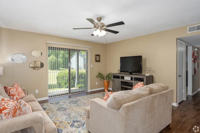 Inviting living room with cozy beige sofas, vibrant orange accent pillows, and an elegant ceiling fan, complemented by a lush garden view through sliding glass doors.