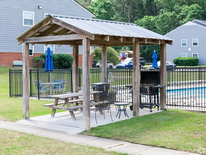 Charming outdoor picnic area with wooden gazebo, barbecue grill, and cozy seating overlooking a refreshing pool.