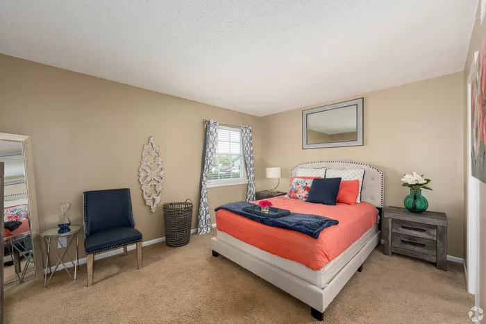 Cozy and well-lit bedroom with a stylish beige palette, featuring a comfortable bed adorned with vibrant orange and floral pillows, a sleek side chair, and elegant decor.