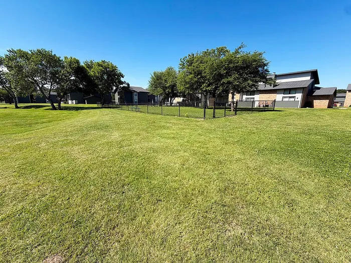 Spacious green lawn with surrounding modern homes and lush trees under a clear blue sky.