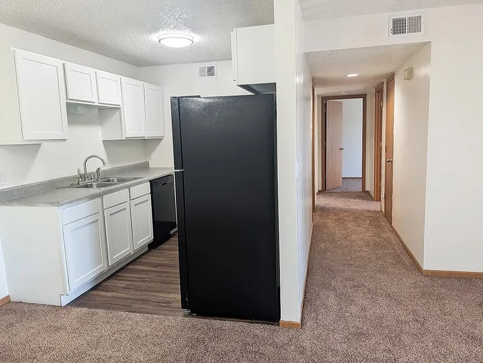 Inviting apartment kitchen with sleek black appliances, ample white cabinetry, and an adjacent cozy carpeted hallway leading to bedrooms.