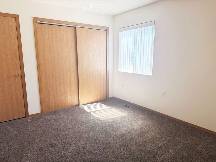 Bright bedroom with soft carpet flooring, large wooden closet, and a sunlit window with blinds.