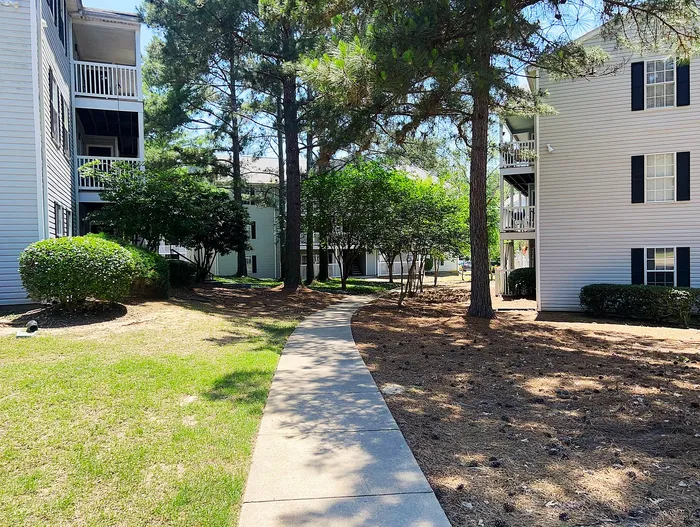 Charming apartment complex exterior with lush greenery and sunlit pathways, offering tranquil living in a serene environment.