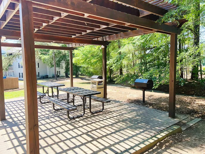Charming outdoor picnic area with wooden pergola, modern tables, and a grill set amid lush greenery.