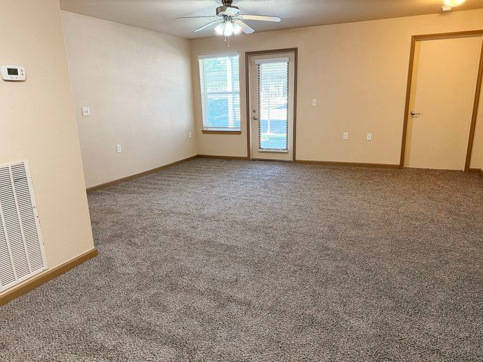 Bright and spacious living area with plush carpeting, modern ceiling fan, and ample natural light from large windows and glass door.
