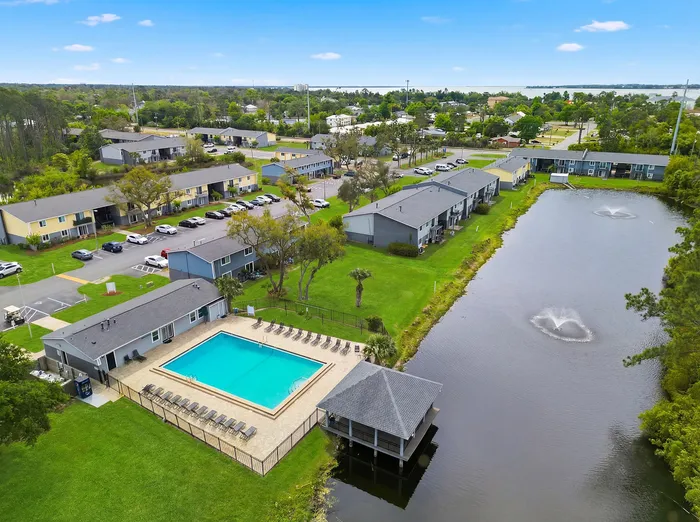 Aerial view of a charming apartment community with a pristine swimming pool, lush green surroundings, and serene pond featuring fountains.