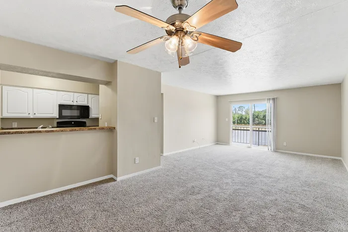 Bright open-plan living room with ceiling fan, adjacent to a modern kitchen with white cabinets and black appliances, featuring large windows with scenic outdoor views.