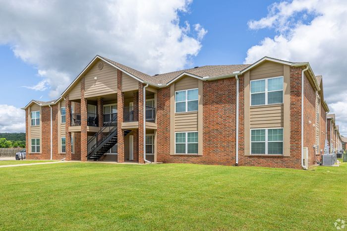 Charming two-story brick apartment building with elegant columns and spacious balconies, set against a vibrant green lawn and a bright blue sky.