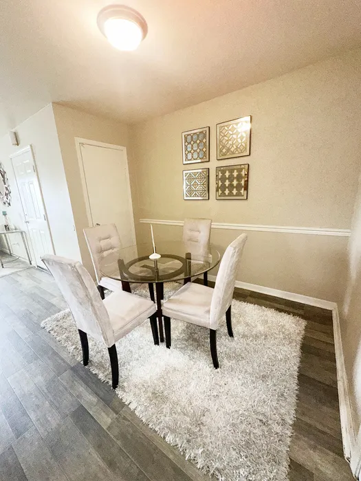 Charming dining area with a sophisticated glass-top table, surrounded by plush beige chairs on a soft white rug, complemented by modern wall art.
