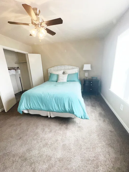Bright and inviting bedroom with plush carpeting, elegant ceiling fan, and comfortable bed with turquoise bedding.