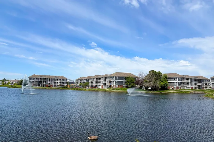 Scenic waterfront apartment complex with elegant buildings overlooking a tranquil lake and fountains, surrounded by lush greenery under a bright blue sky.