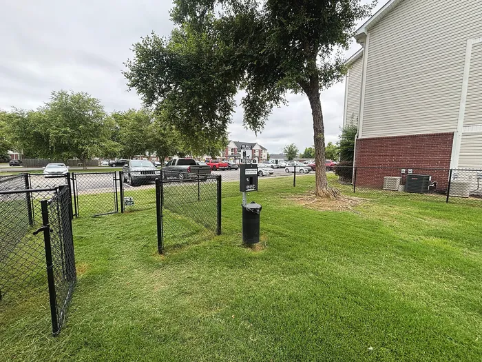 Charming apartment exterior featuring a well-maintained grassy area with pet-friendly fenced section, mature trees, and ample parking space.