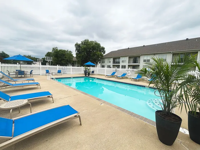 Inviting apartment community pool area with vibrant blue loungers, shaded seating, and lush greenery, offering a relaxing getaway.