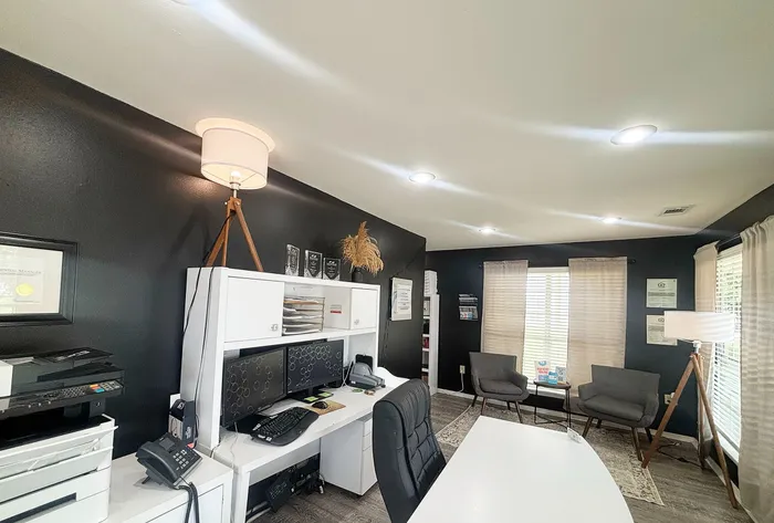 Chic and modern office space with sleek black walls, elegant white furniture, dual monitors, and cozy seating area near large sunlit windows.