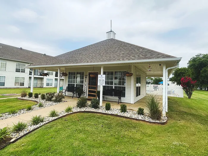 Charming outdoor view of a welcoming apartment entrance with lush green lawn, decorative landscaping, and inviting porch seating.