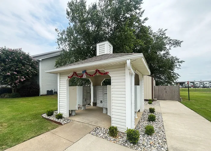 Charming white gazebo-style mailbox pavilion with patriotic decorations, surrounded by lush greenery and a serene walkway.