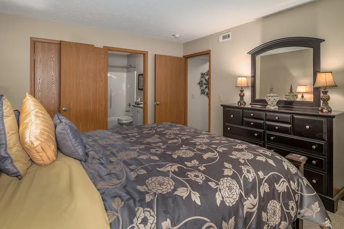 Inviting bedroom with plush floral bedding, wooden dresser with elegant lamps, and convenient en-suite bathroom.