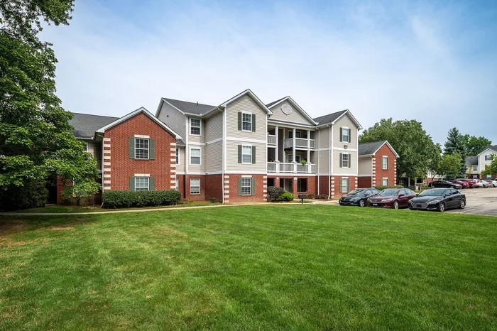 Charming apartment complex featuring brick accents, lush green lawn, and ample parking space against a bright, clear sky.